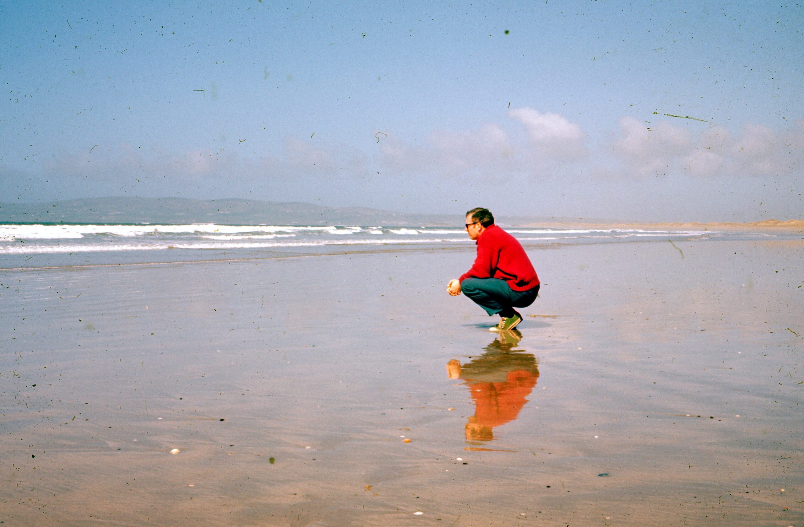 A Man Kneeling on the Sandy Beach, Gazing at the Ocean Waves Under a Clear Blue Sky.
