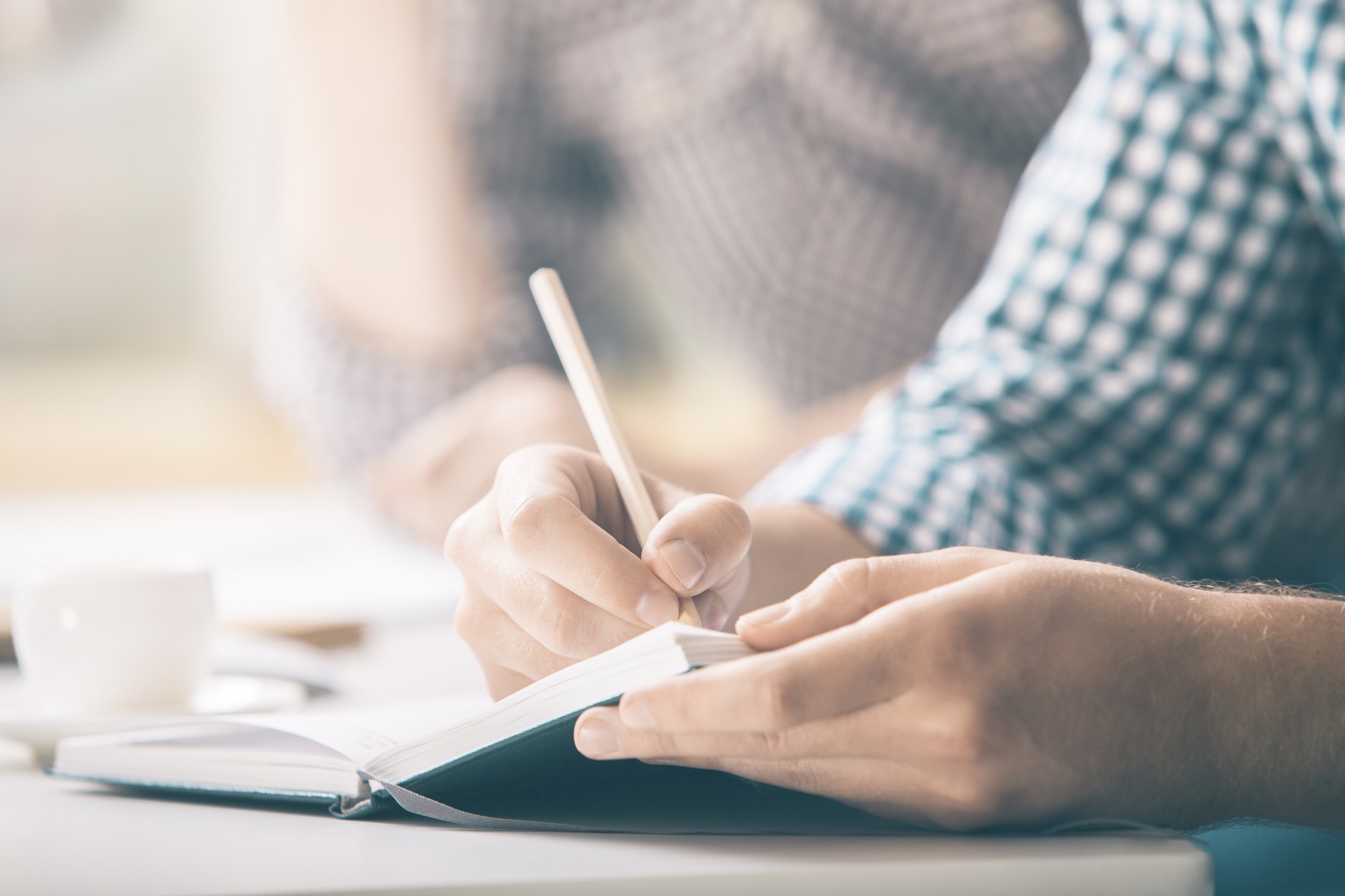 Male Writing in Notepad Placed on Desktop With Coffee Cup, Supplies, Equipment and Other Items. Close Up